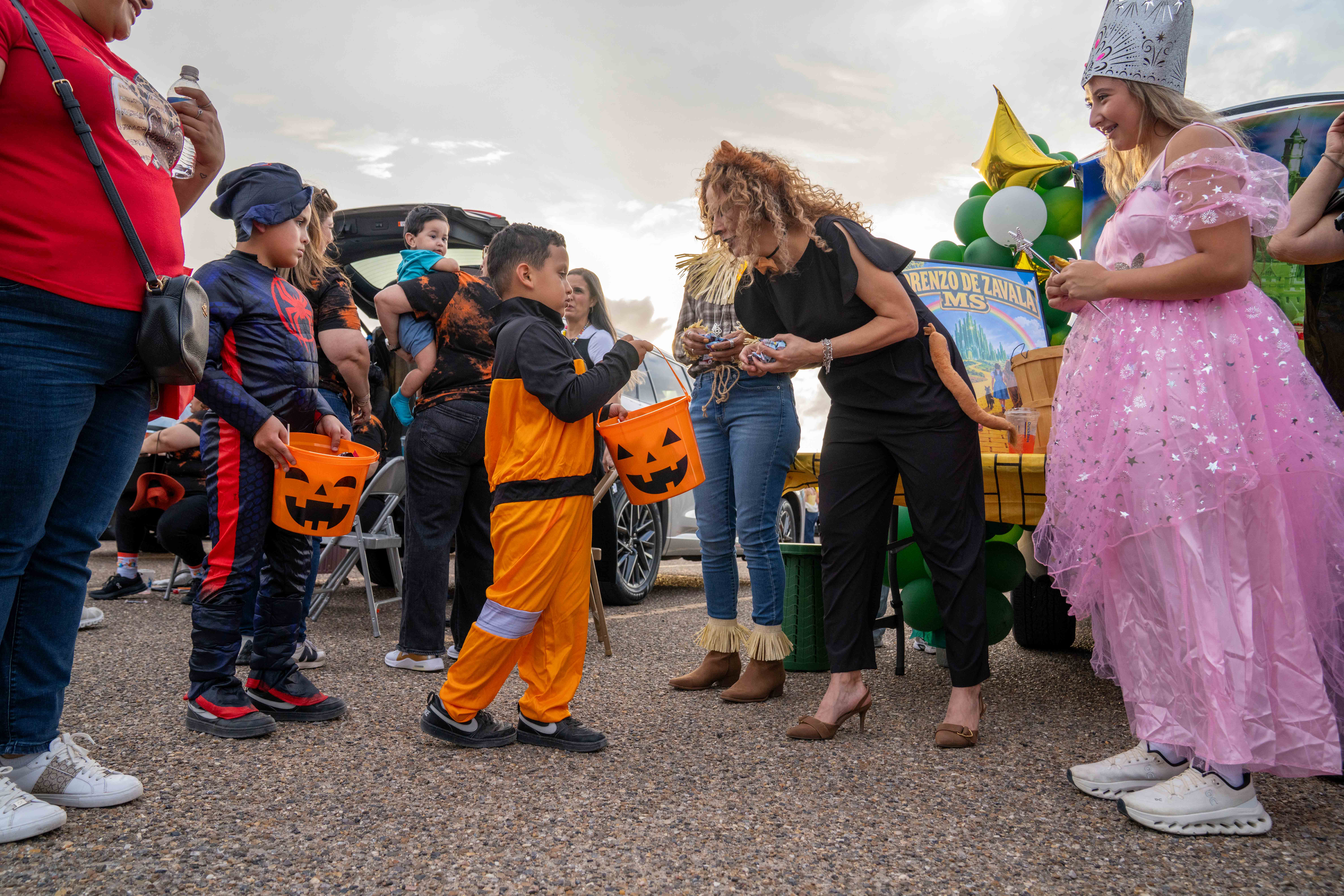 Children in Halloween costumes collect candy from adults during a daytime trunk-or-treat event outdoors.
