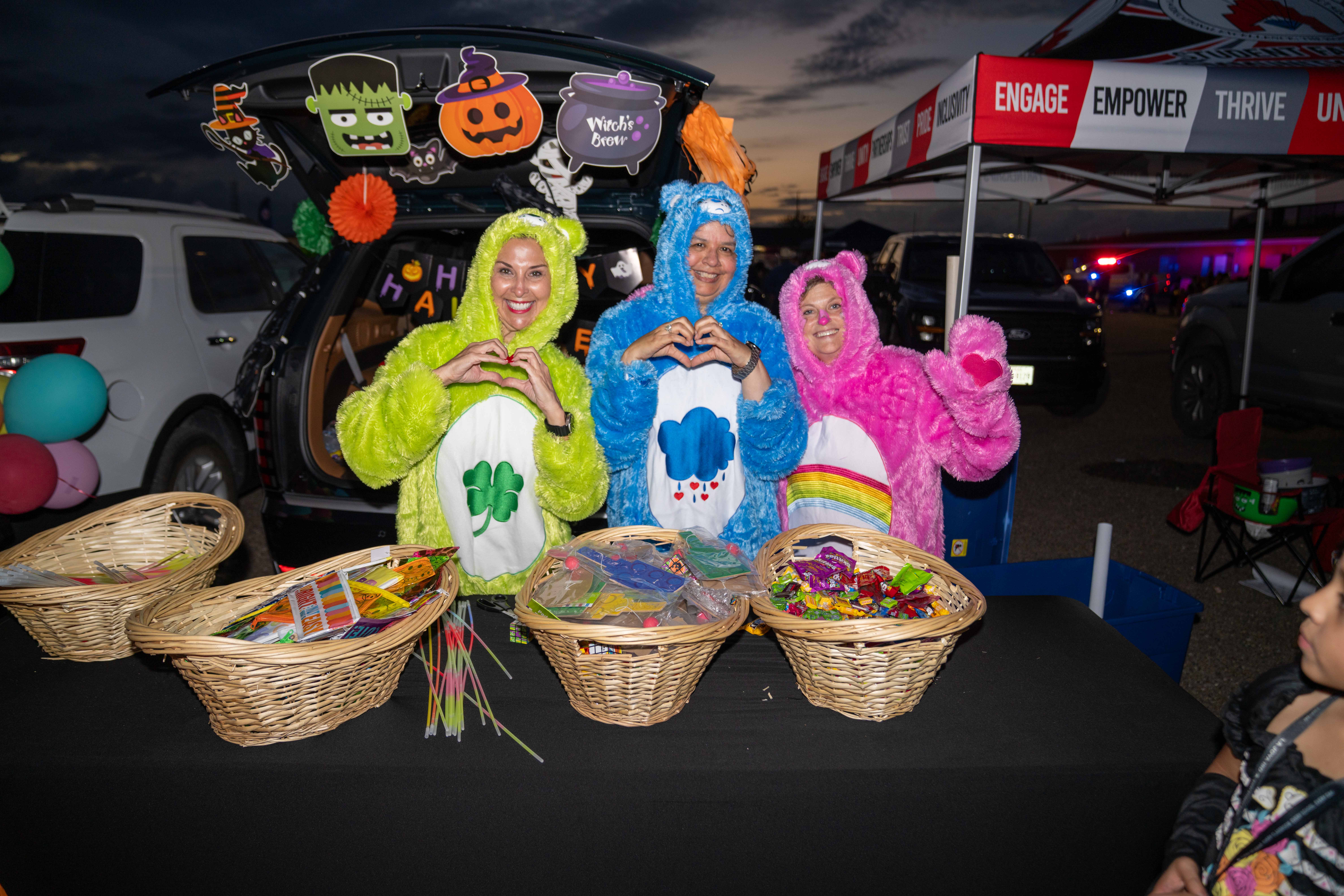 Three people in colorful bear costumes pose behind a table filled with baskets of candy and glow sticks at a trunk-or-treat event.