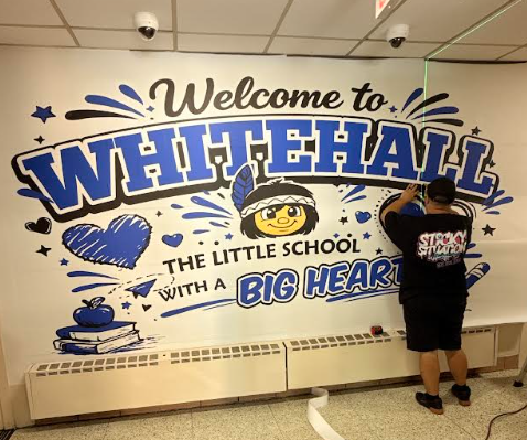 A person is installing a large vinyl wall mural that reads "Welcome to WHITEHALL THE LITTLE SCHOOL WITH A BIG HEART."