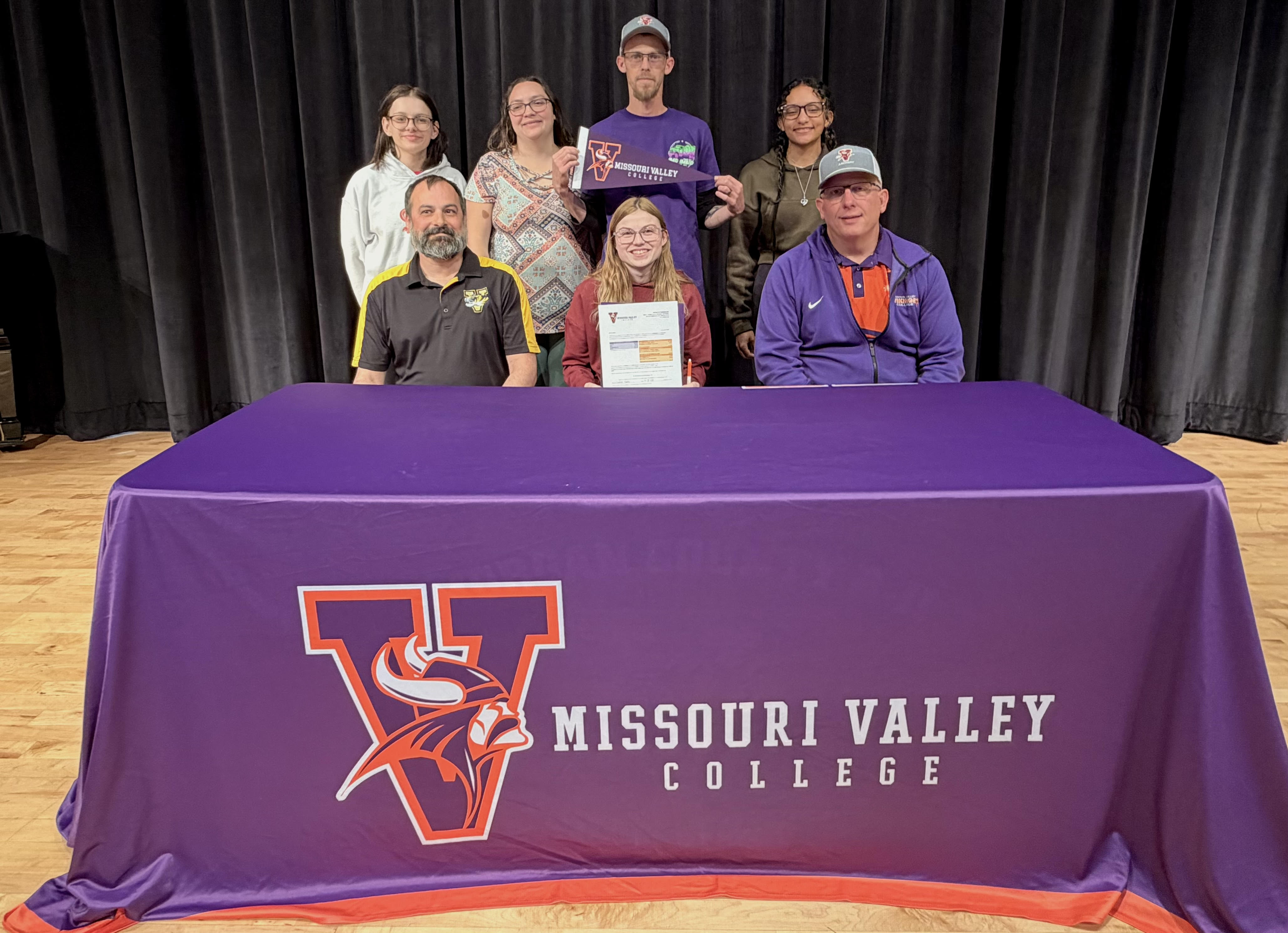 A group of people, including students and adults, pose for a photo in front of a black curtain for a college signing for archery.