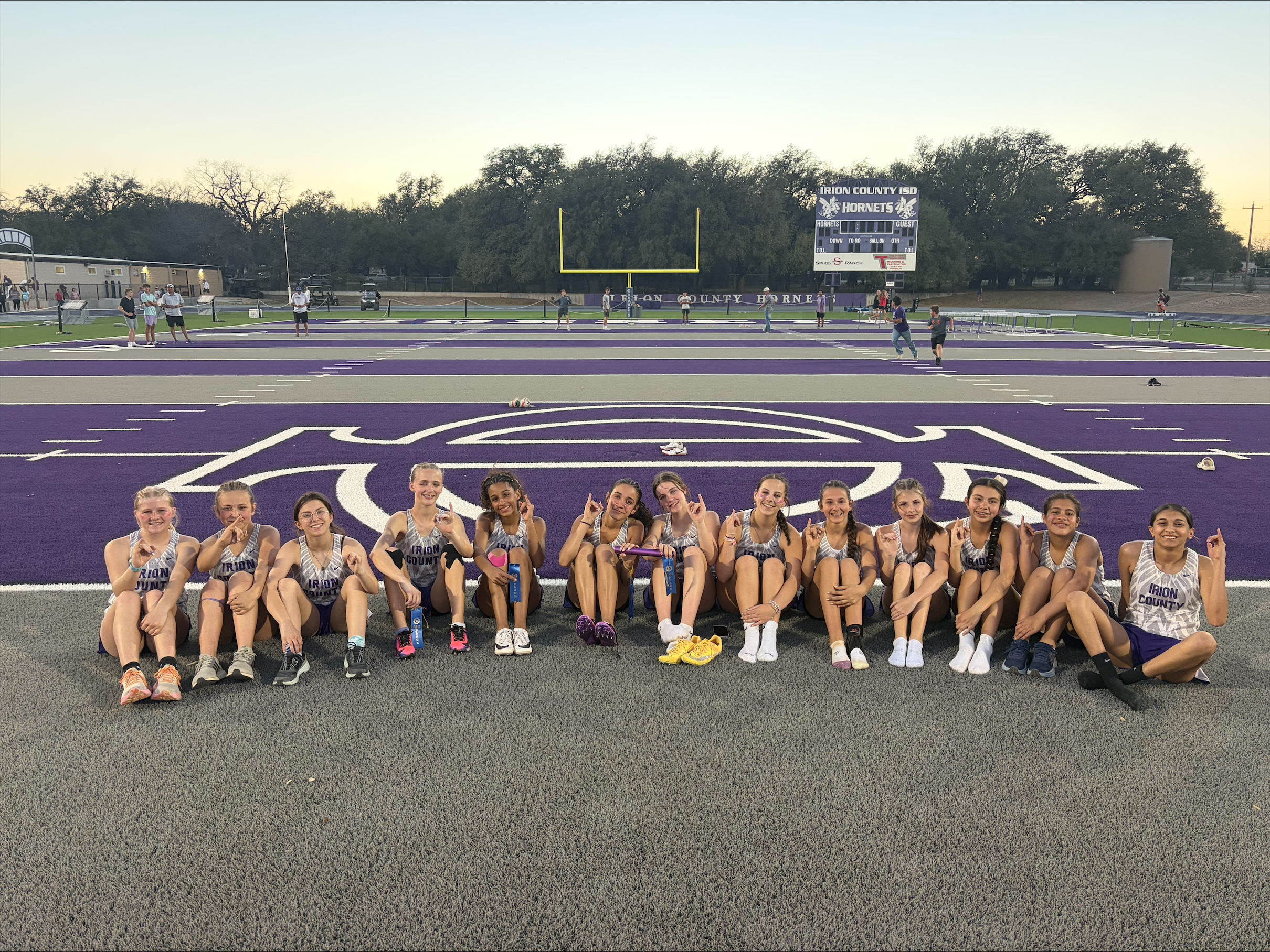 A group of young women in track uniforms sit on a track field, smiling and posing for a photo.