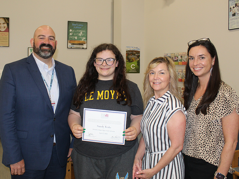A young woman receives a certificate from three adults in an indoor setting.