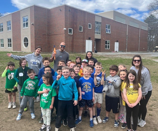 A group of smiling children and adults pose for a photo in front of a brick school building.
