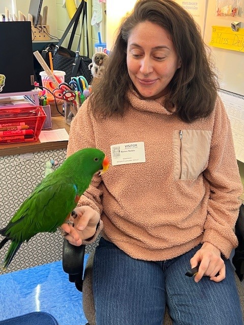 A woman wearing a visitor badge smiles as a green parrot perches on her hand.
