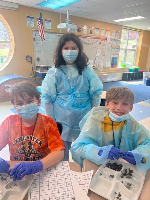 Three students in a classroom wear protective gowns and masks, engaged in a science activity.