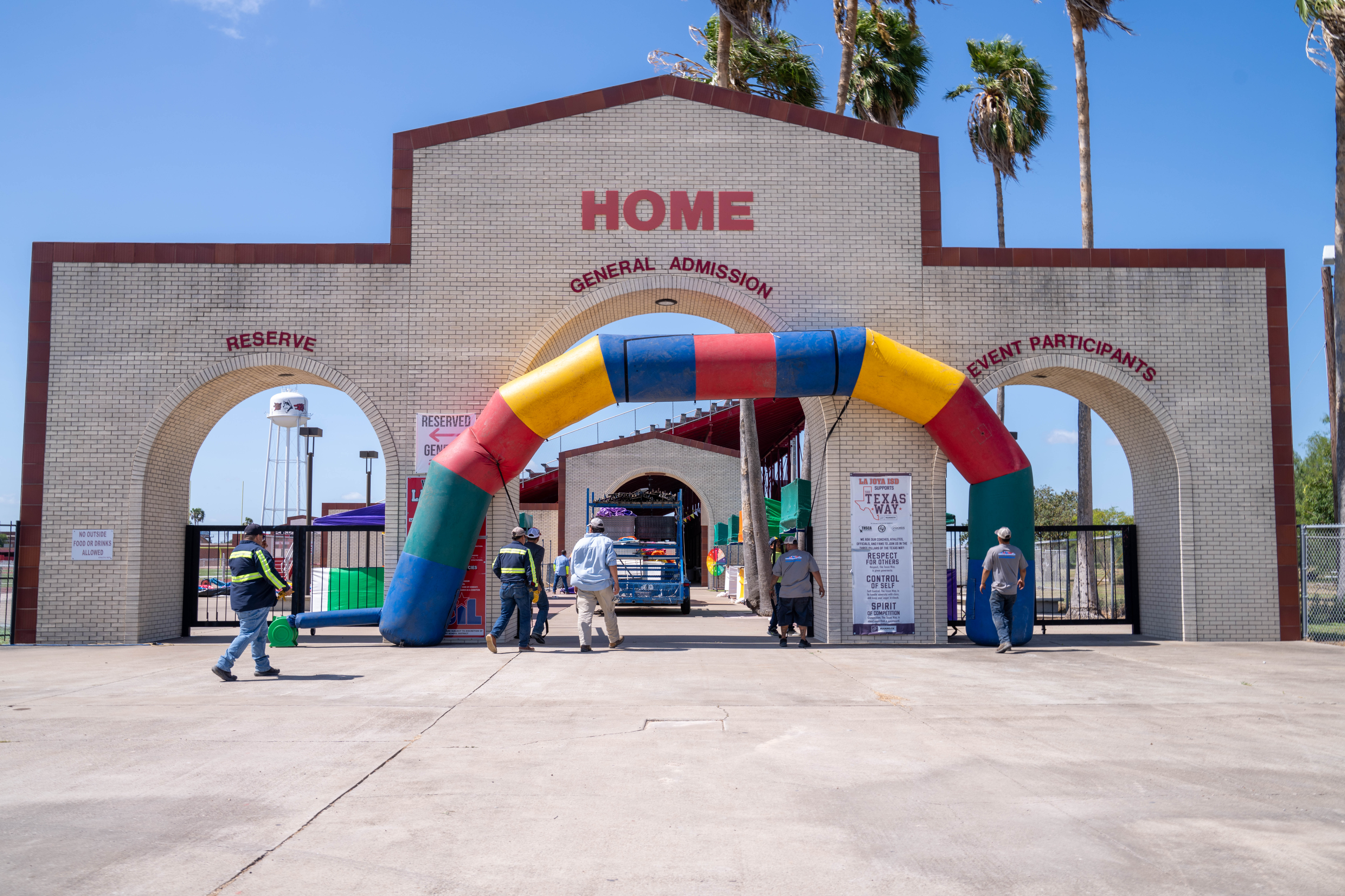People walk under a colorful inflatable archway at the entrance to a venue with signs for 'HOME', 'GENERAL ADMISSION', 'RESERVE', and 'EVENT PARTICIPANTS'.
