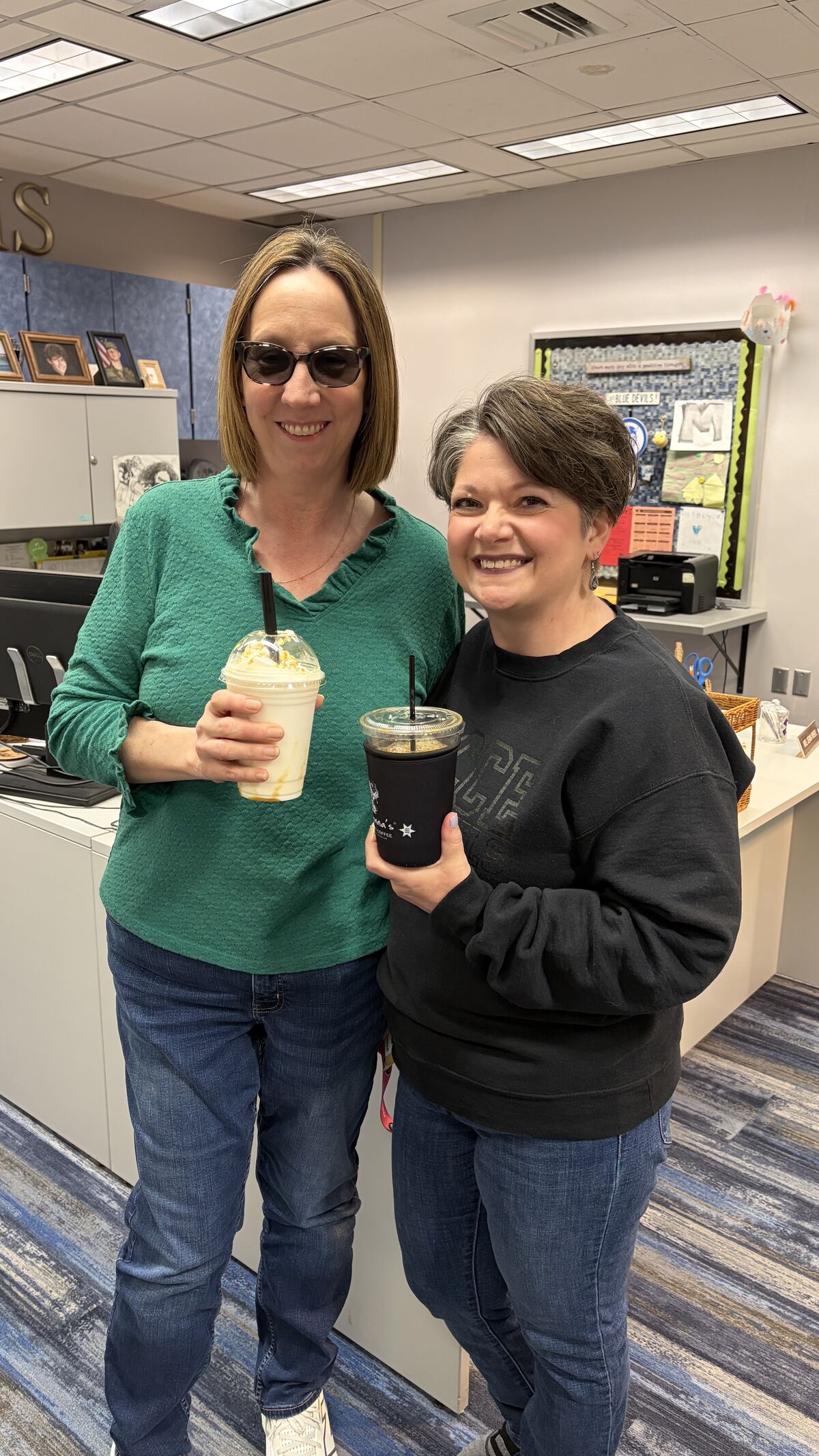 Two women smile while holding drinks in an office setting.