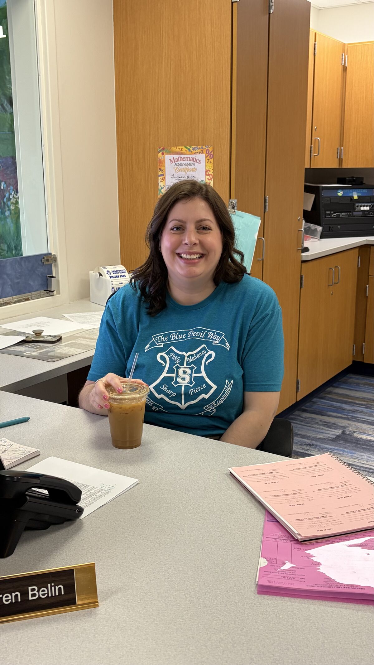 A woman with dark hair smiles while holding a coffee drink at a desk.