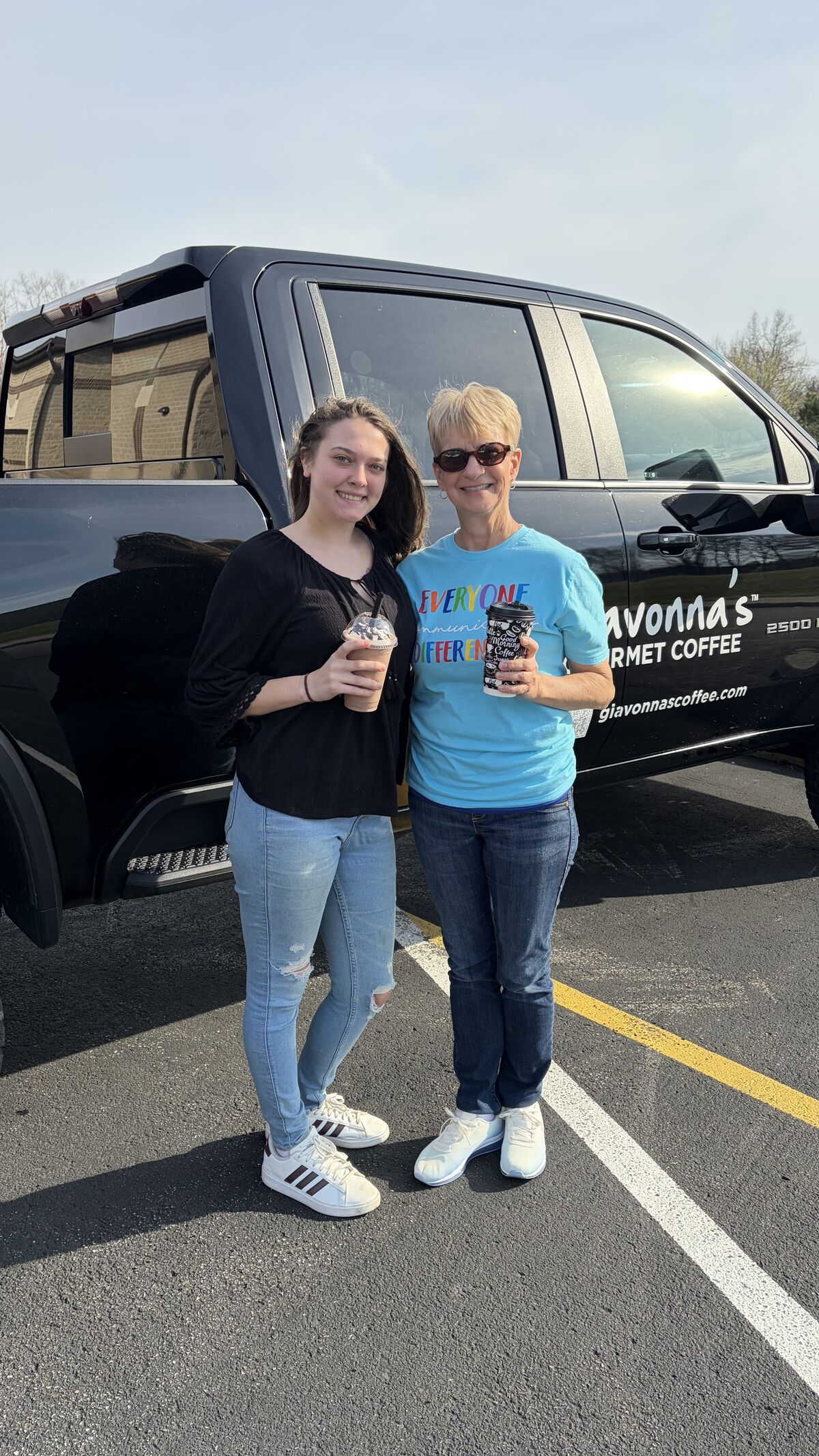 Two women stand in front of a black truck, each holding a coffee cup.