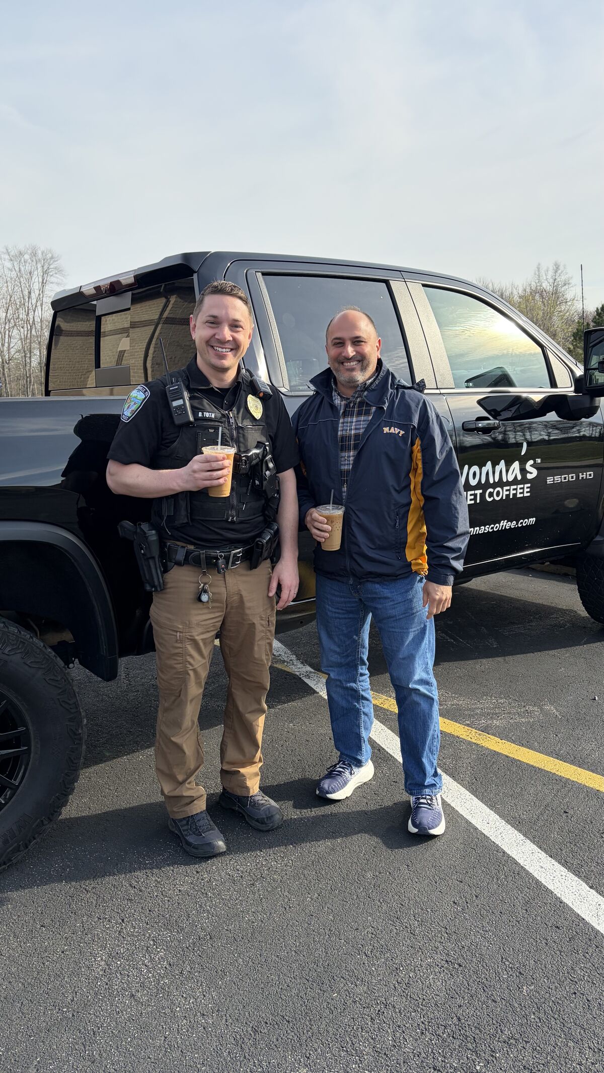 A police officer and a civilian stand next to a black pickup truck, both holding iced coffee drinks.