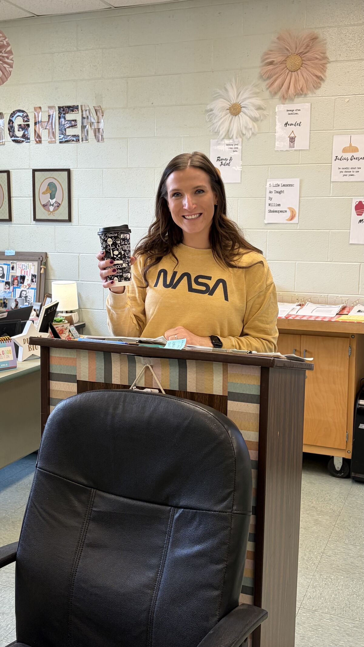 A woman in a yellow NASA sweatshirt smiles and holds a coffee cup in an office setting.
