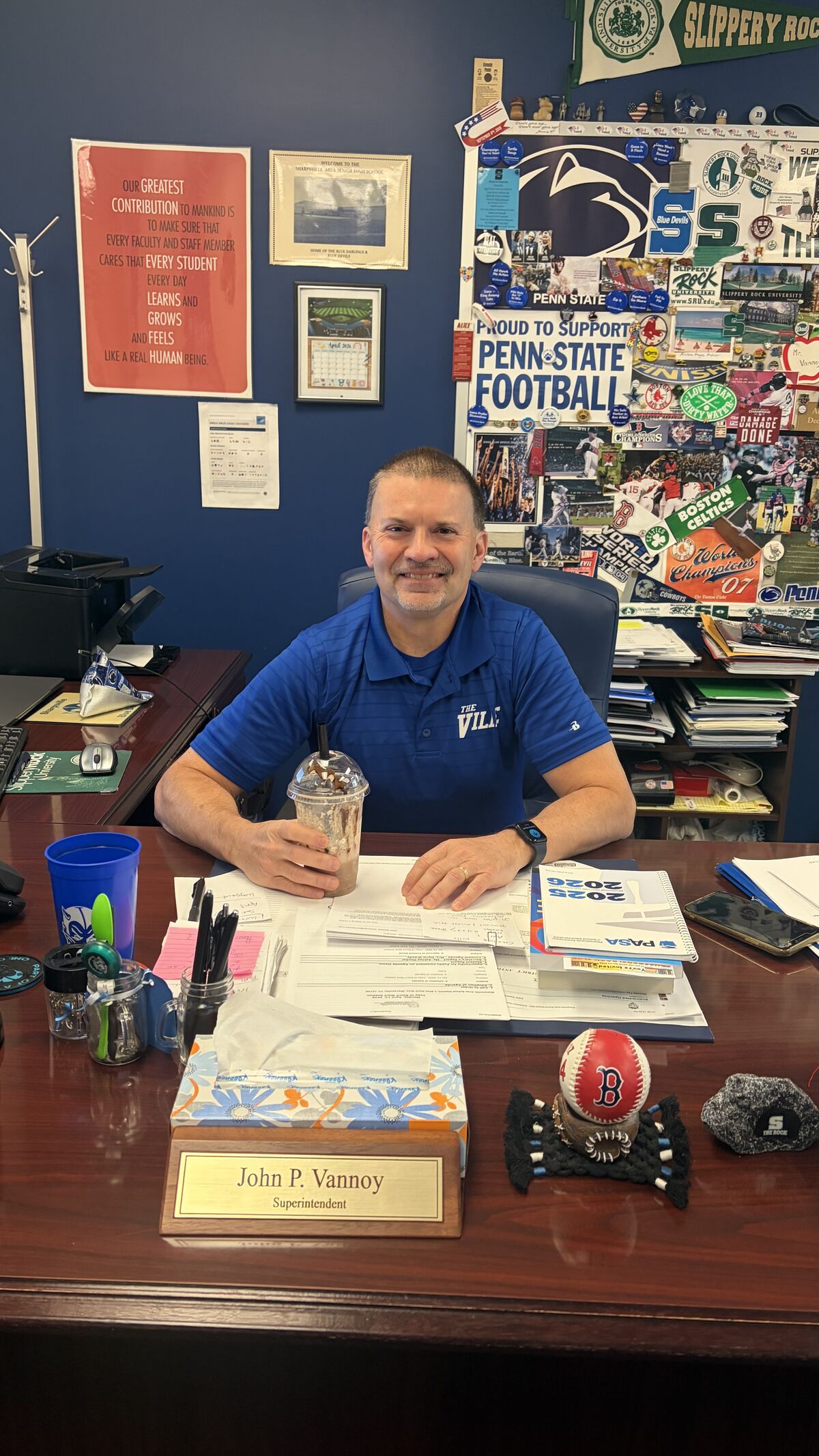 A smiling man in a blue polo shirt sits at a desk in an office decorated with sports memorabilia and motivational posters.