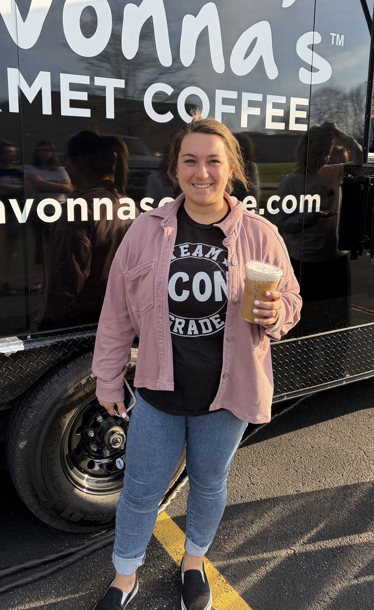 A smiling woman holds a large iced coffee in front of a black coffee truck with white lettering.