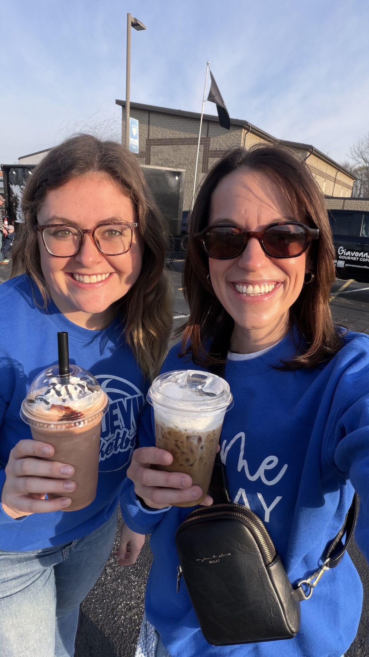 Two smiling women in blue sweatshirts hold iced coffee drinks outdoors.