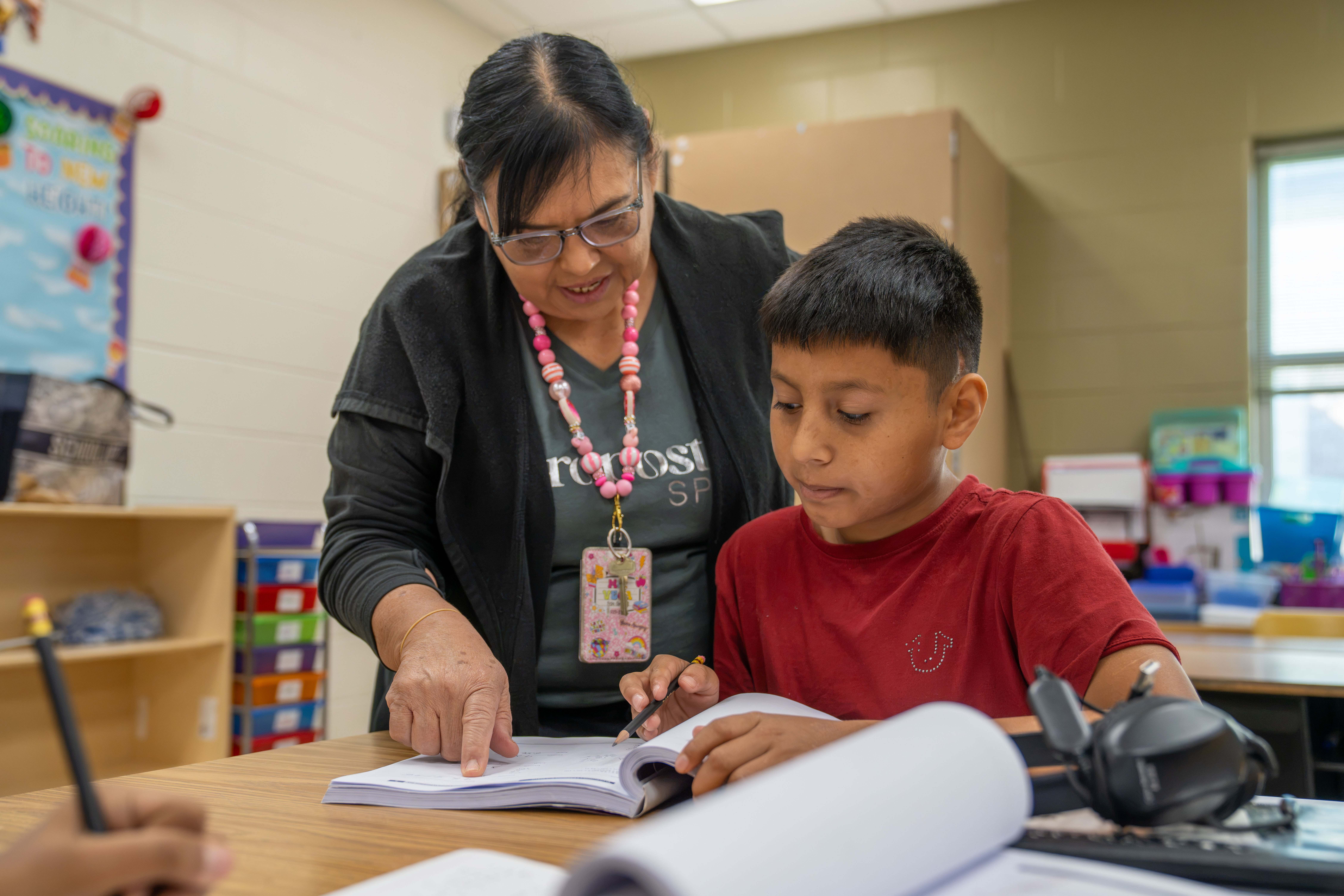 A teacher guides a young student as they work together on a book at a desk in a classroom.