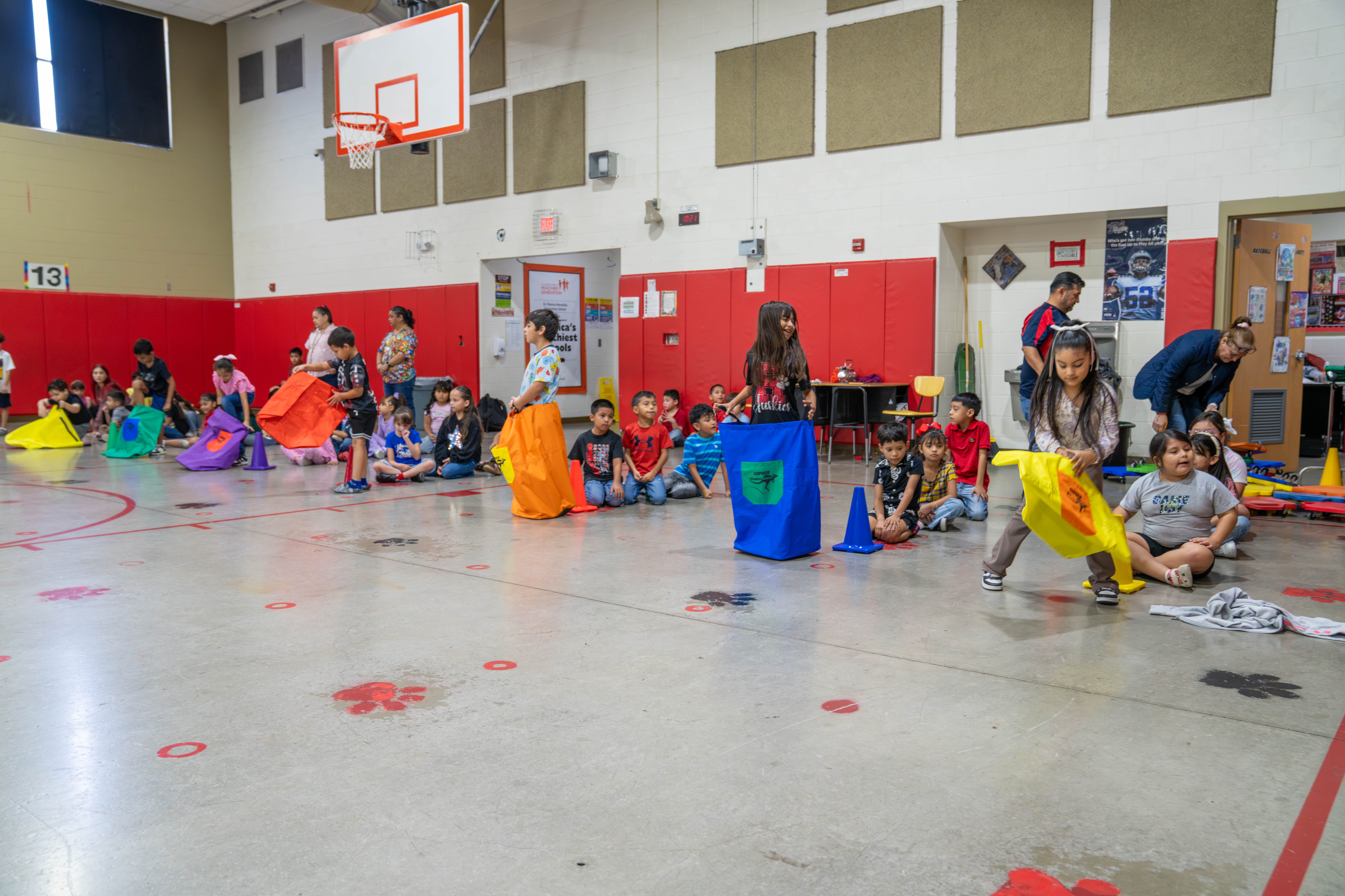 Children participate in a sack race in a school gymnasium with colorful sacks and cones.
