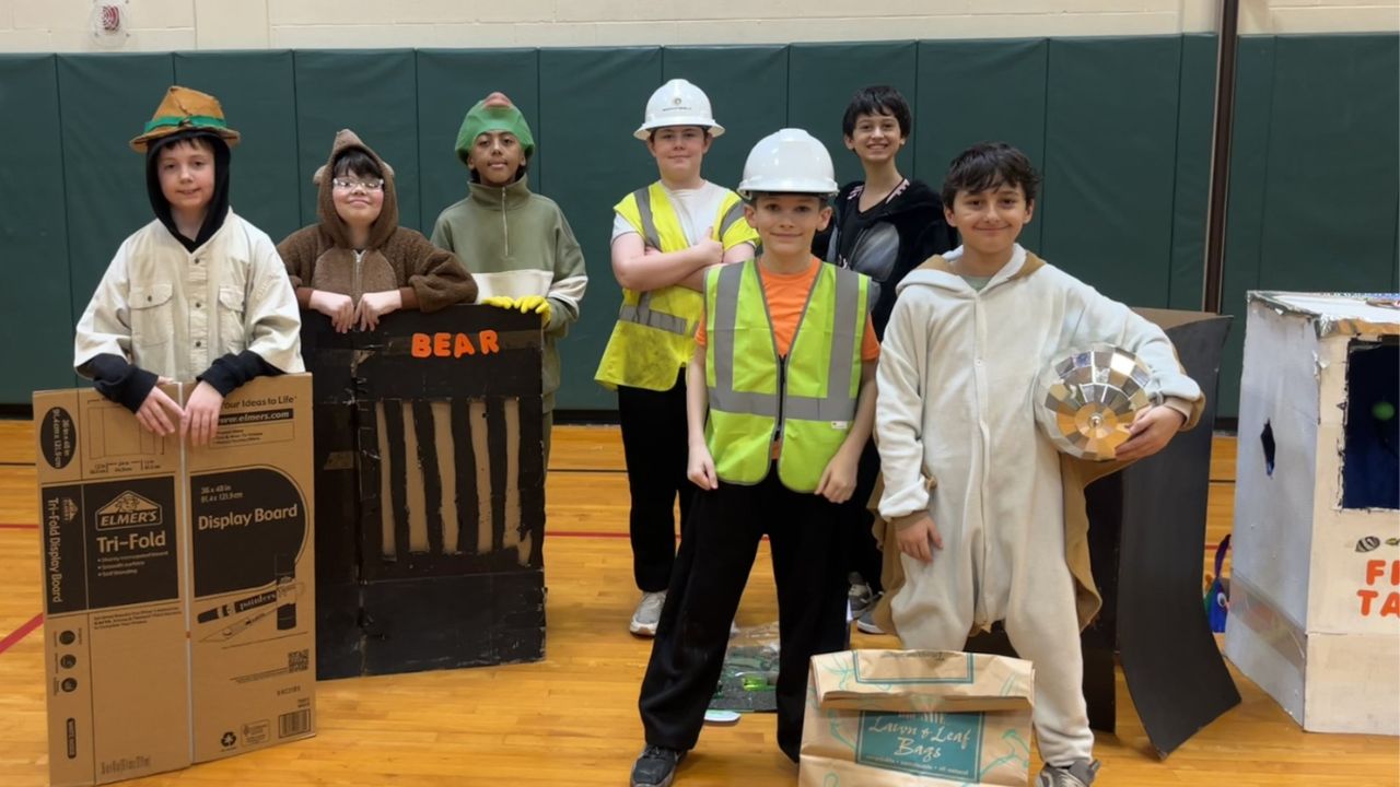 A group of children in costumes and safety vests stand in a gymnasium, posing with cardboard props.