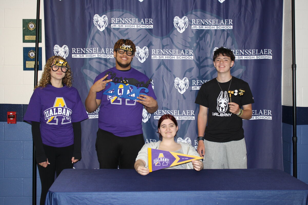 Four students pose for a photo in front of a Rensselaer Jr-Sr High School banner, holding graduation-themed props.
