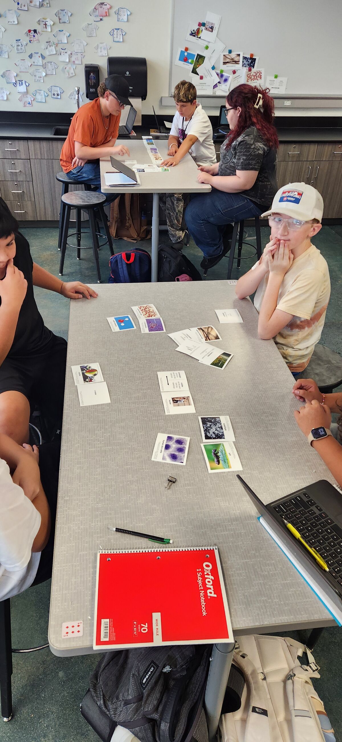 Students gather around a table in a classroom, looking at educational cards and a laptop.