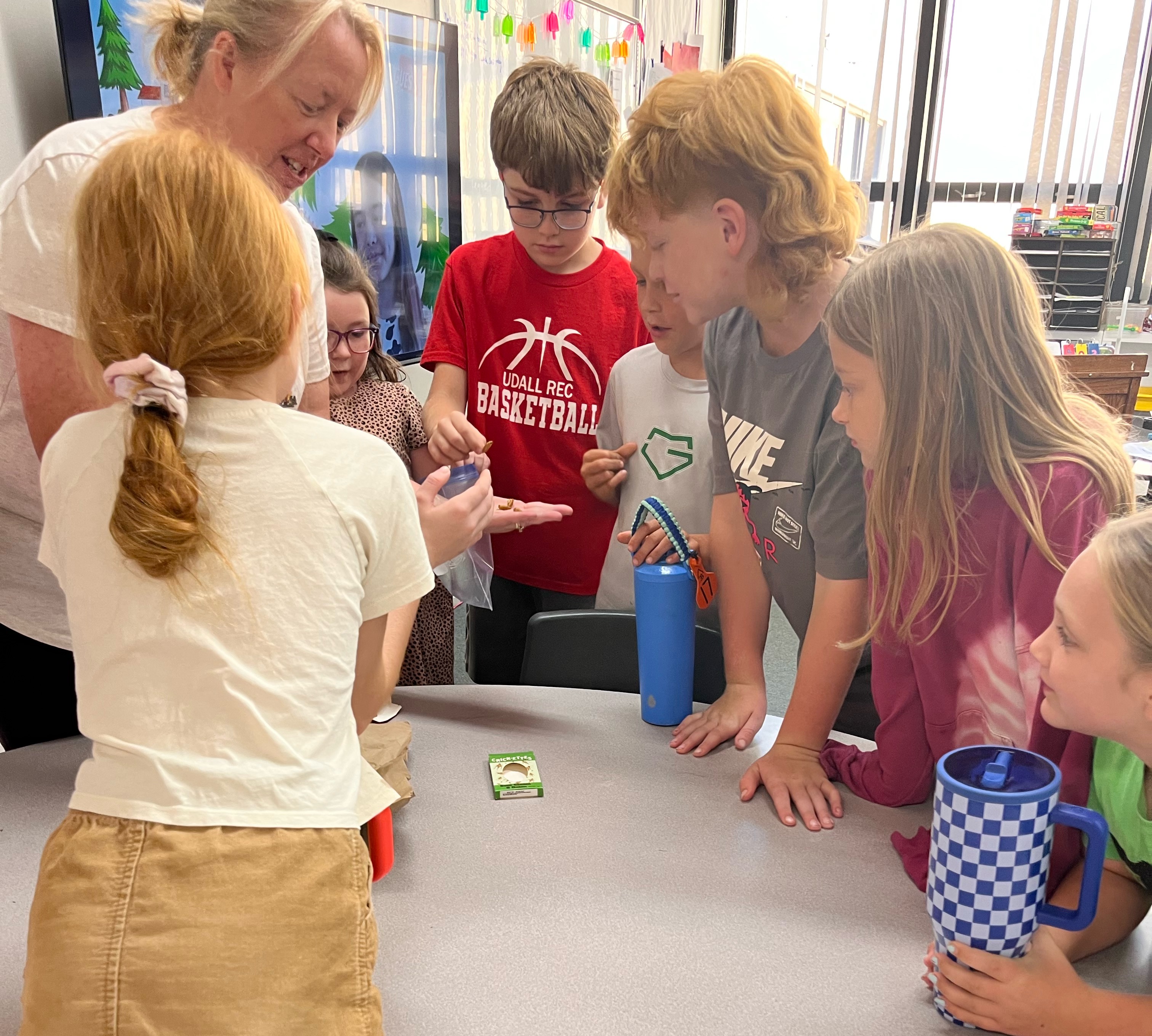 A teacher and a group of students gather around a table in a classroom, looking intently at something.
