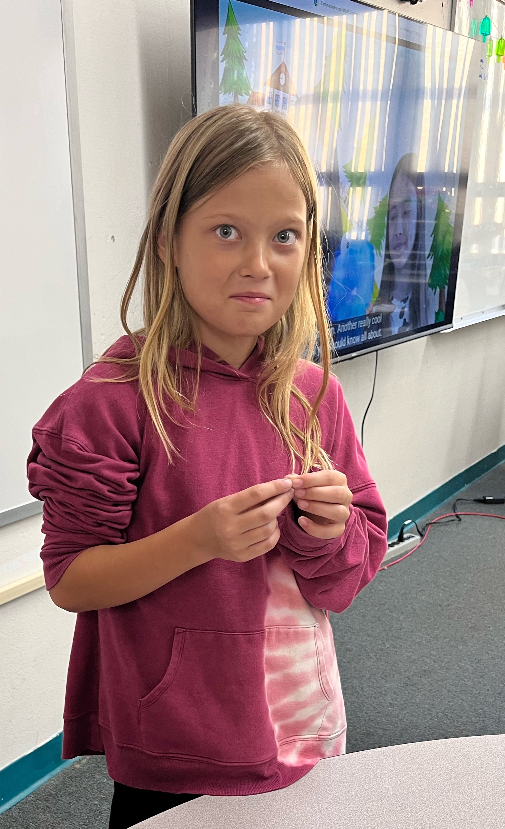 A young child with long blonde hair and a maroon hoodie stands in a brightly lit classroom.