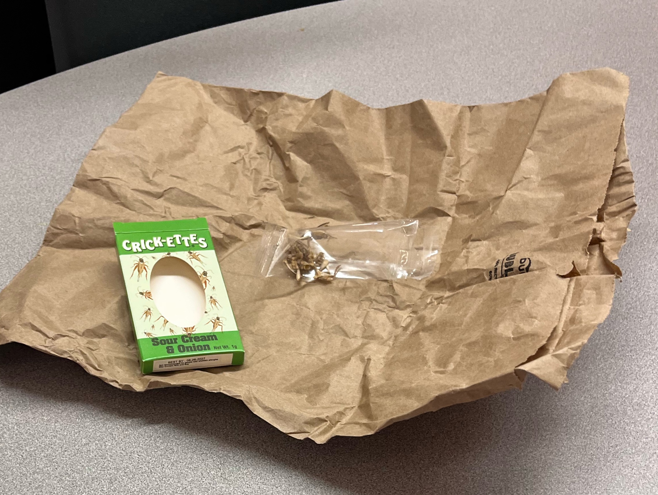 A box of Sour Cream & Onion flavored Crick-Ettes sits on a crumpled brown paper bag on a table.