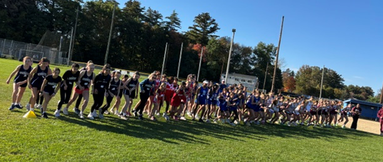A large group of runners lines up on a grassy field to start a cross-country race on a sunny day.