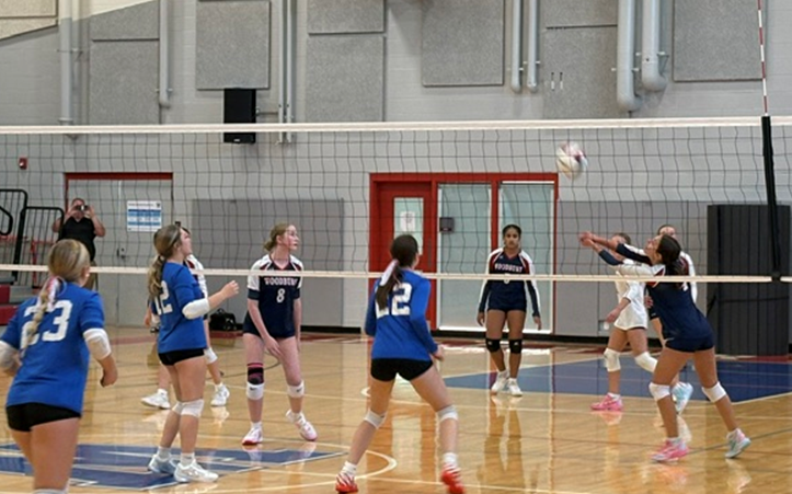 A girls' indoor volleyball team plays a match in a gymnasium, with players in blue and dark jerseys focused on the ball near the net.