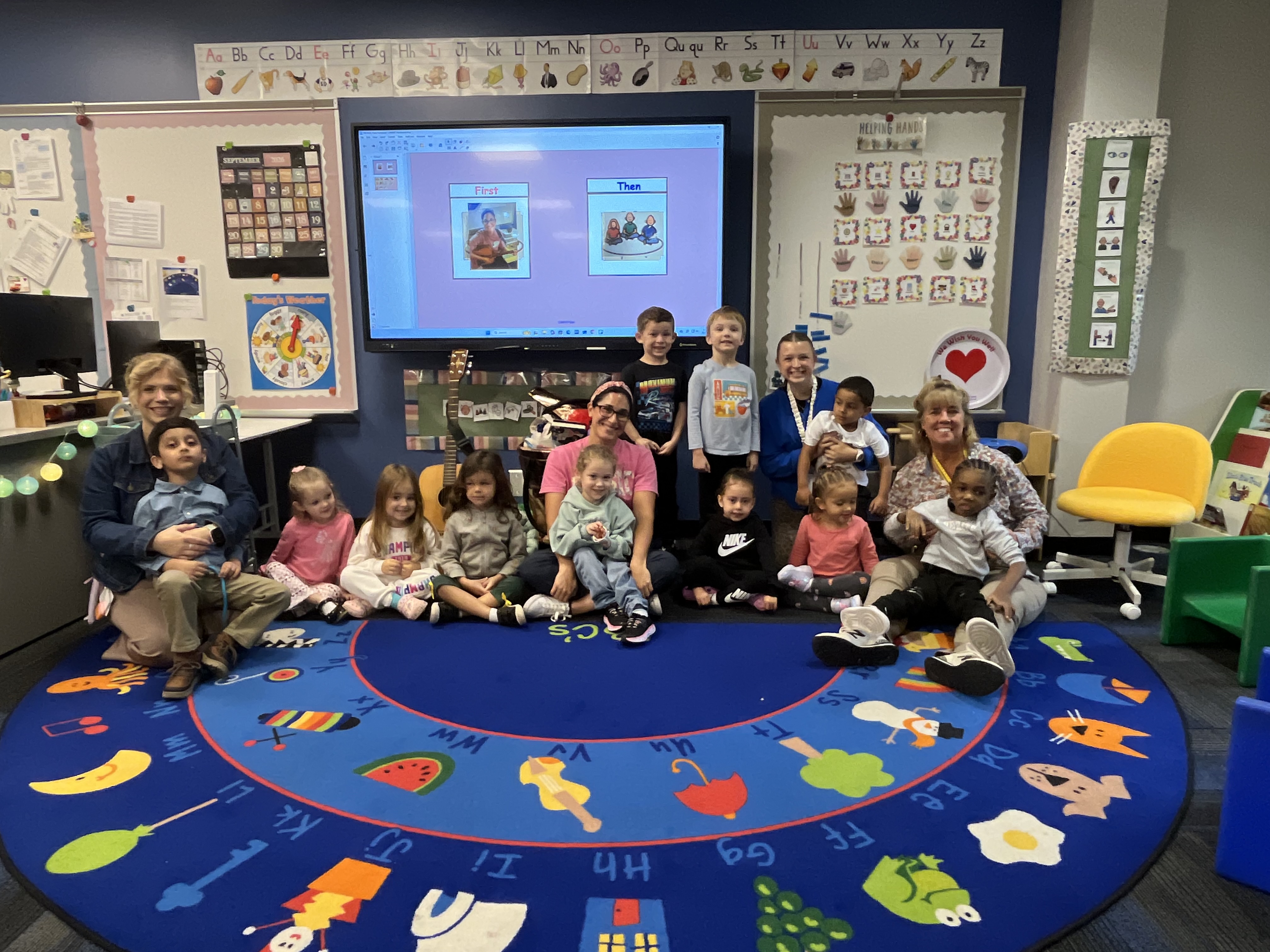 Miss Abbey, teachers and students smile in a group photo while seated on the carpet