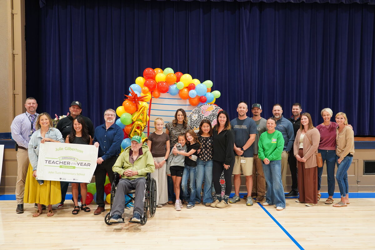 A diverse group of adults and children pose for a photo on a school stage in front of a colorful balloon arch and a blue curtain.