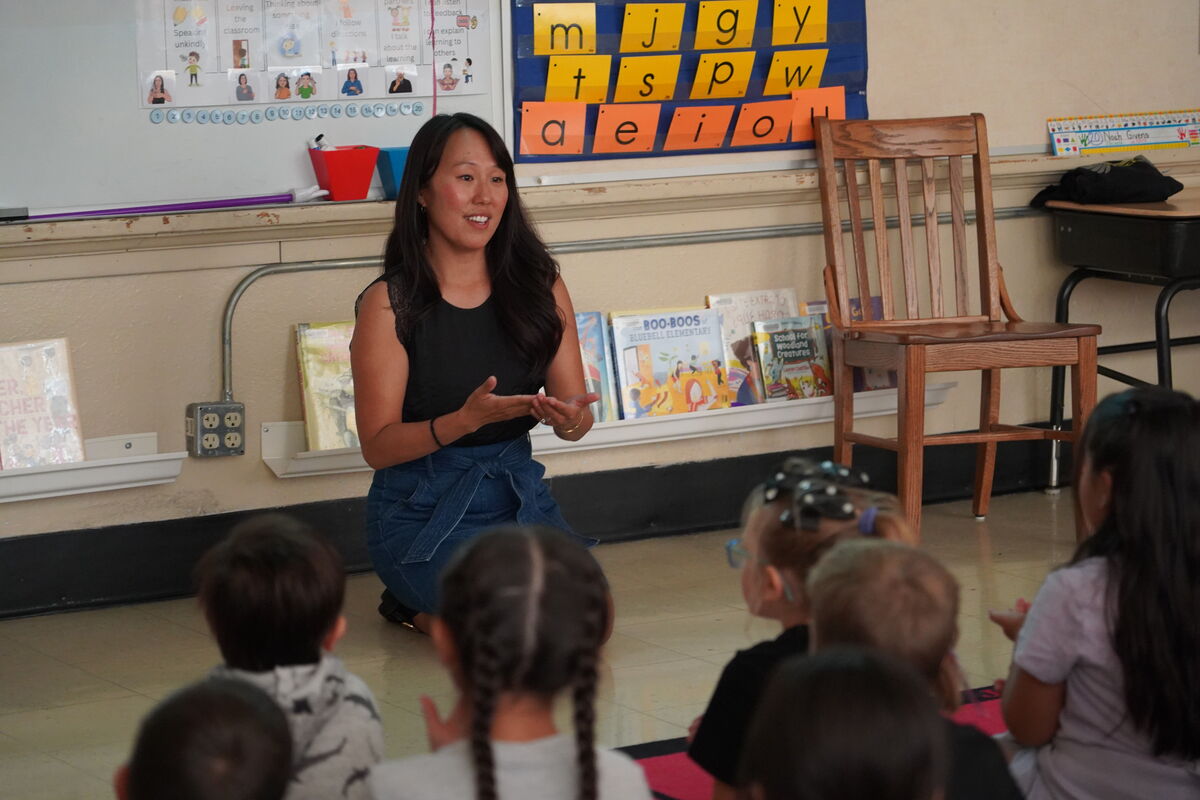 A teacher kneels on the floor, smiling and gesturing as she talks to a group of young students sitting in front of her in a brightly lit classroom.