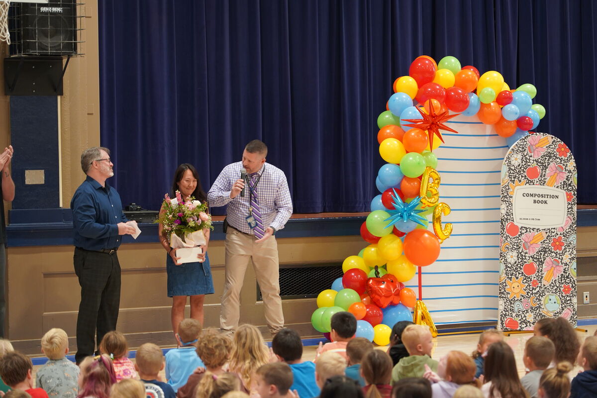 A man speaks into a microphone on a stage while another man applauds and a woman holds a bouquet of flowers.