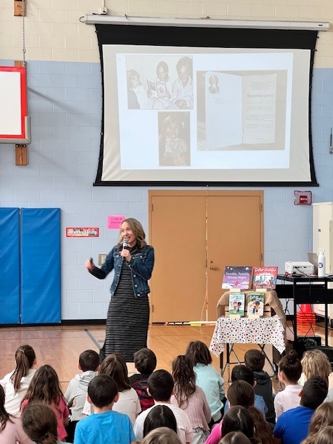 A woman with a microphone speaks to a group of children sitting on the floor in front of a projector screen.