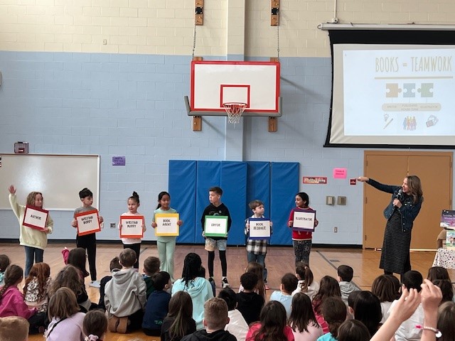 Children hold signs for 'Author', 'Writing', 'Book Agent', and 'Illustrator' during a school presentation.