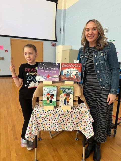 A young girl in a 'READ LEAD' shirt points to books displayed on a table.