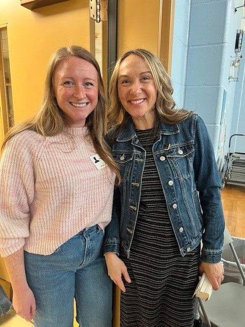 Two smiling women stand side-by-side, posing for a photo indoors.