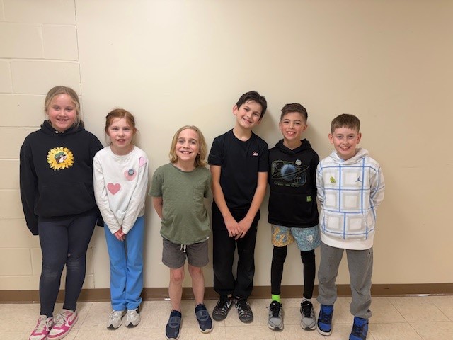 Six smiling children stand in a line against a light-colored wall.