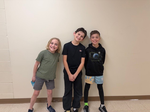 Three smiling boys stand together against a light-colored wall.