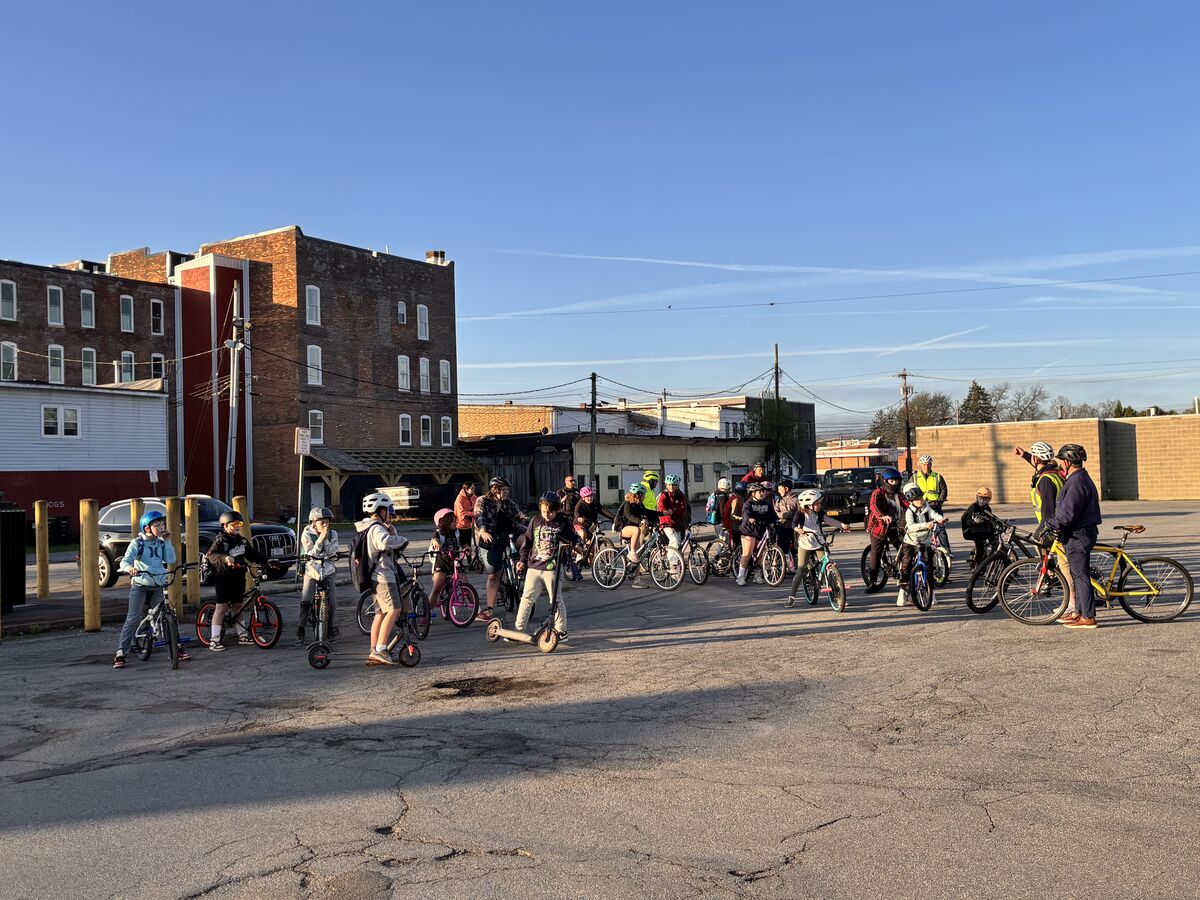 A diverse group of people, including children and adults, gather with bicycles and scooters.