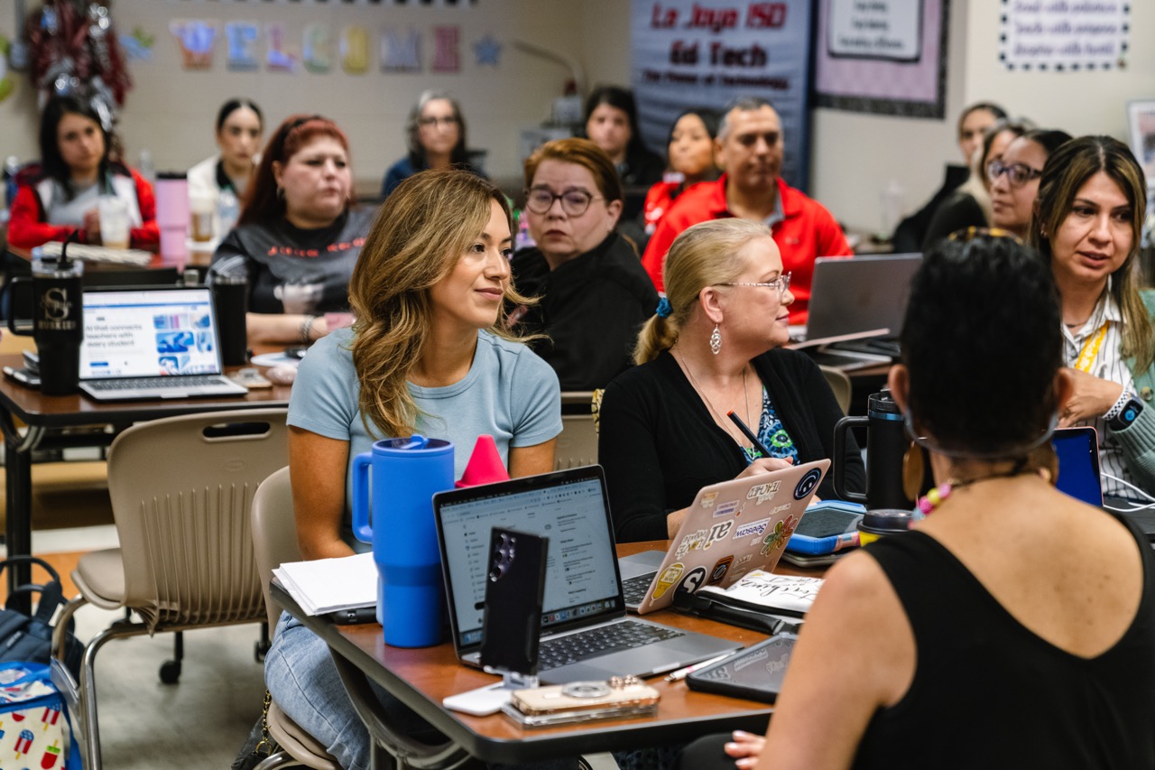 A group of adults attend a professional development session in a classroom, with laptops and notebooks on their desks.