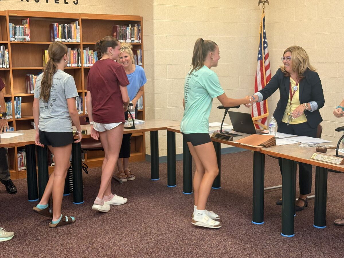 A woman in a suit shakes hands with a young woman in a teal t-shirt in a room with bookshelves.