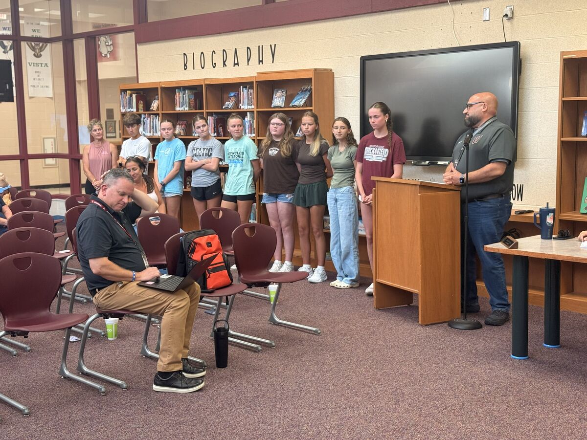 A group of students stands in front of a podium in a library, with a teacher speaking.