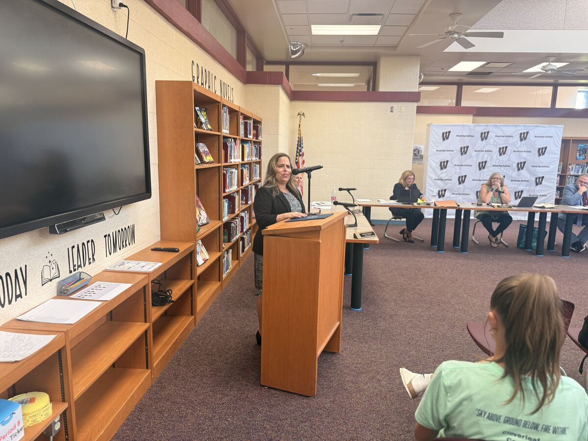 A woman speaks at a podium in front of bookshelves and an audience in a library setting.
