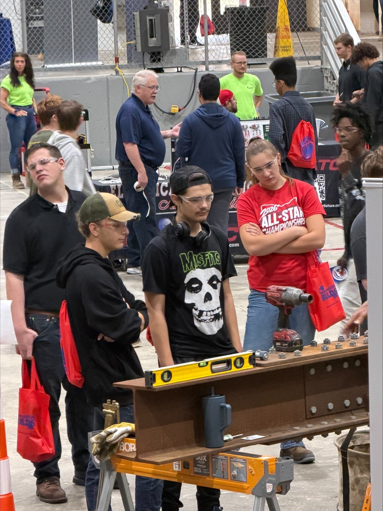 A group of students wearing safety glasses observes a structural beam exhibit at an indoor venue.