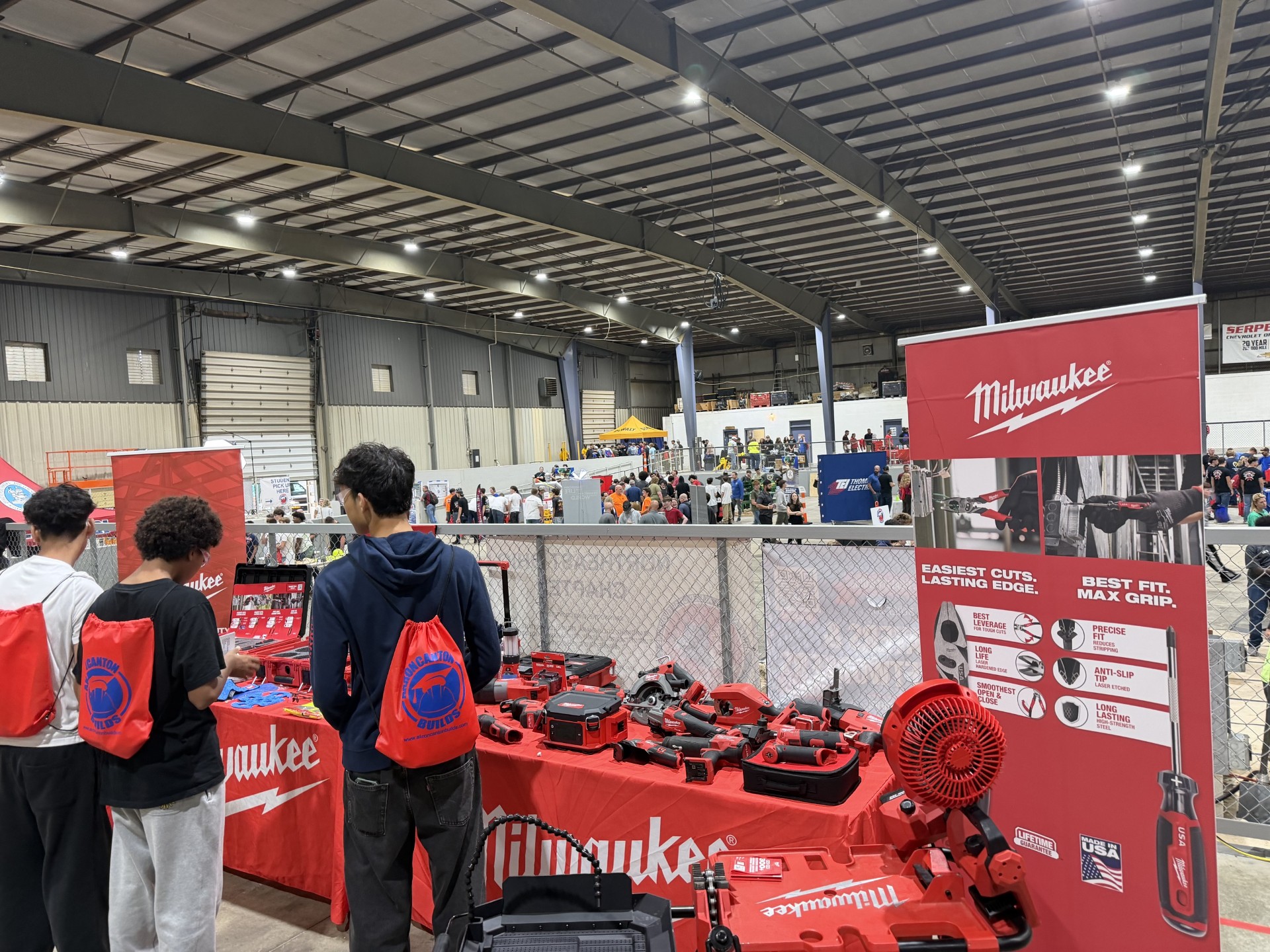 Three people look at Milwaukee tool displays at a trade show inside a large warehouse.