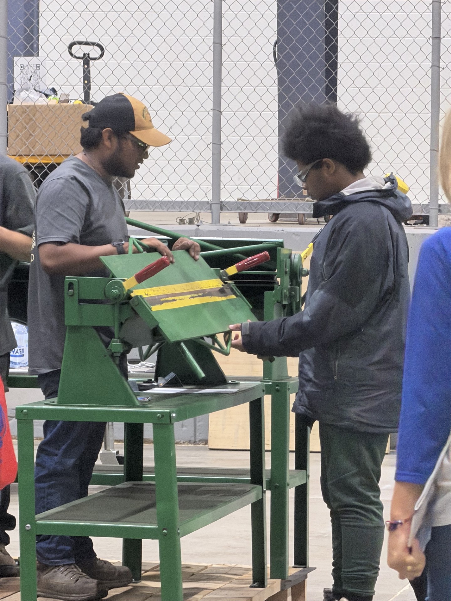 Two people collaborate to operate a green metal-bending machine in an industrial workshop.