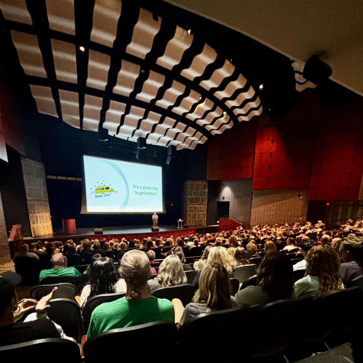 An audience watches a presentation on a large screen in a theater.