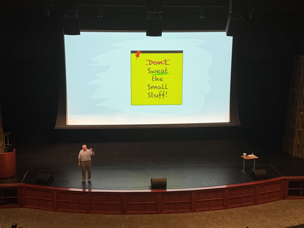 A speaker stands on a stage in front of a large screen displaying a yellow sticky note with the text 'Don't Sweat the Small Stuff!'.