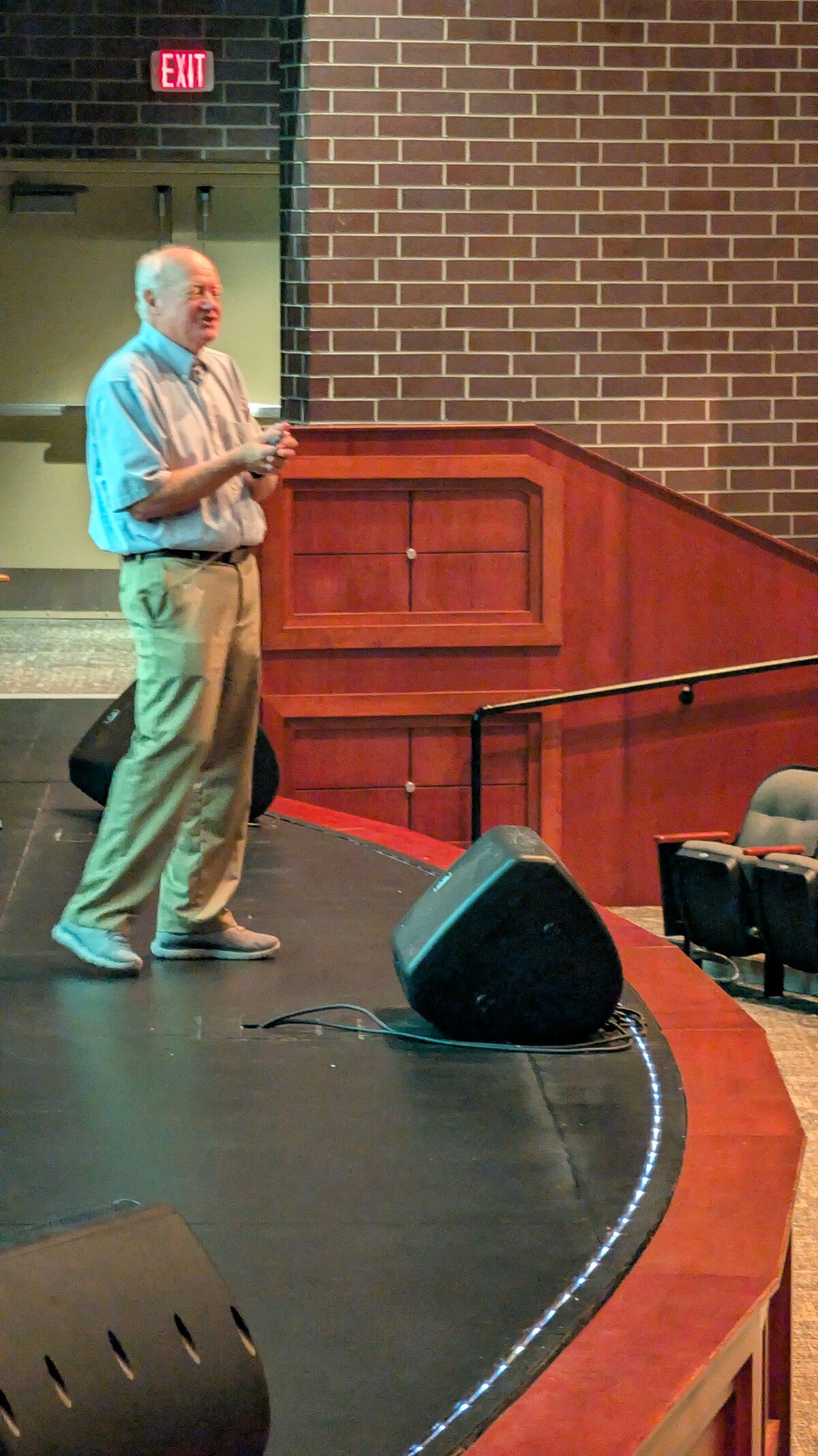 An older man in a light blue shirt and khaki pants stands on a stage, holding a microphone.