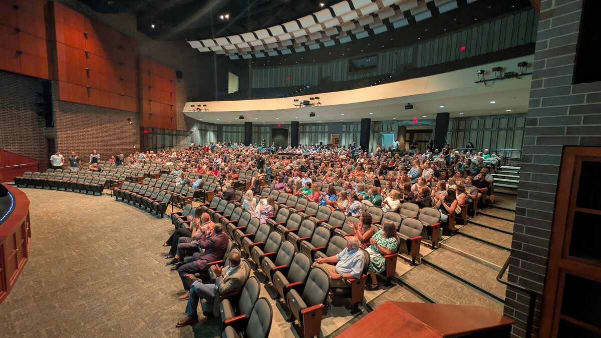 A large audience fills a tiered auditorium, seated in rows of dark chairs.